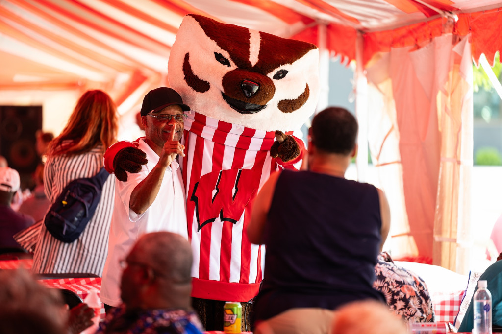 Under a red and white event tent, a man holds up his finger to make a No. 1 sign while posing with Bucky Badger.