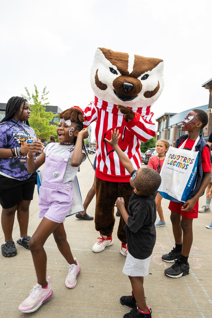 A group of children laugh and dance with Bucky Badger at a community picnic.