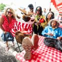 A group of women in colorful blouses gather around a seated Bucky Badger at a picnic table with a red and white checked table cloth. Bucky is sitting with his feet up on the table as everyone around him laughs and smiles.