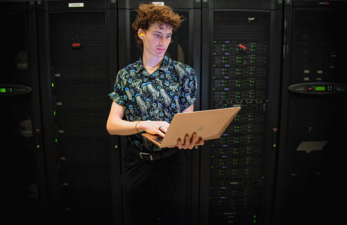 A portrait photo of Grant Nickle, wearing a shirt with a vibrant blue jellyfish pattern, stands holding a laptop computer in a server room.