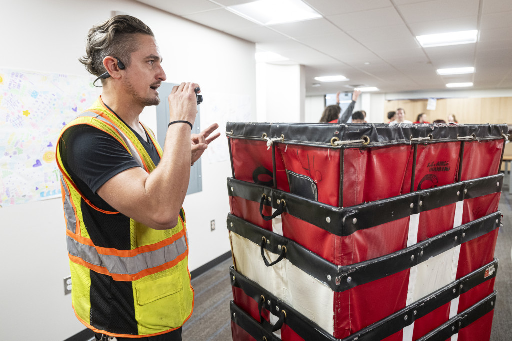 A man stands in a hallway. He is wearing a yellow safety vest and talking into a walkie-talkie. In front of him is a stack of large, red industrial laundry bins on wheels. 