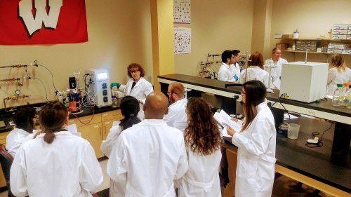 In a laboratory, people in white coats gather around a woman operating a piece of equipment. A UW-Madison flag hangs on the wall.