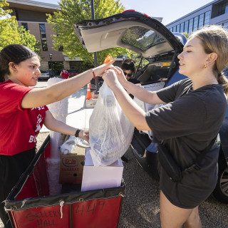 A woman in a red shirt helps a woman load belongings from the back of a car into a red cart.