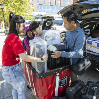 A family works together to load belongings from the back of a car into a red cart.