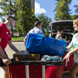 A man in a red shirt steadies a red moving cart while a woman places a blue duffle bag inside.