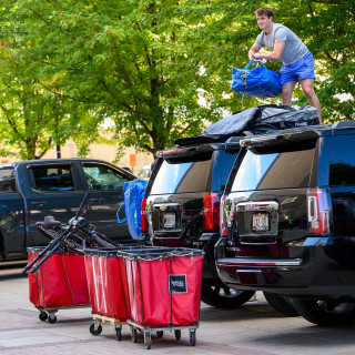 A new student stands on top of an SUV and tosses a blue duffle bag into a red cart.