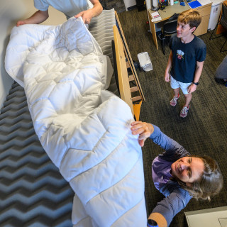 A woman reaches up to spread a comforter across the top bunk in her son's dorm room.