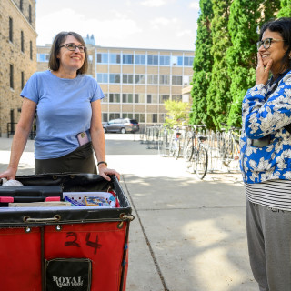 Two moms talk over a red moving cart outside a residence hall.