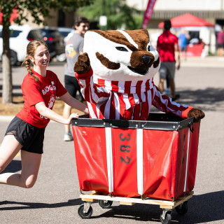 Bucky Badger rides inside a red cart pushed by a Badger Buddy.