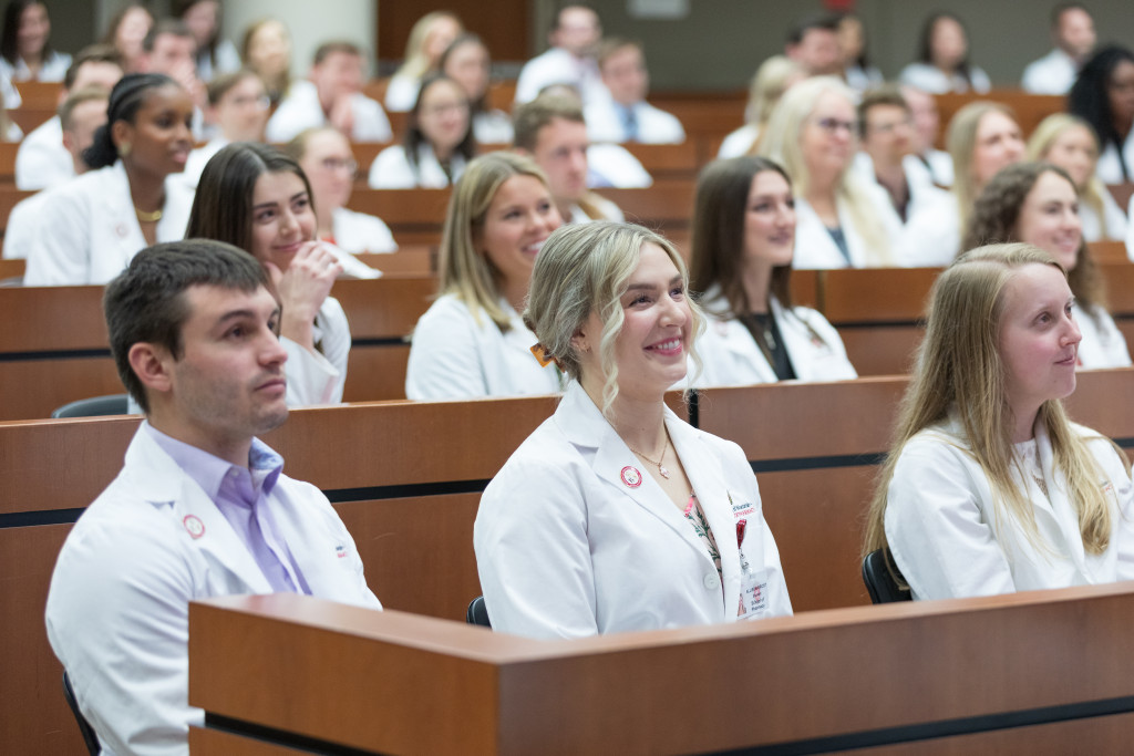 A group of students wearing white coats sit in a hall.