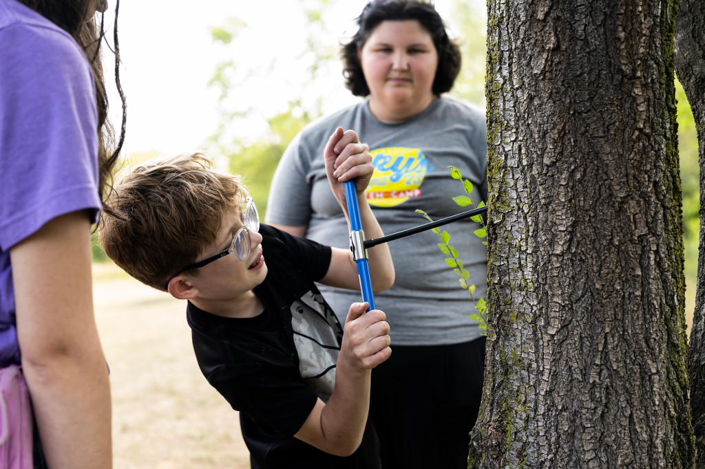A boy uses a tool to drill into a tree trunk as a woman watches.