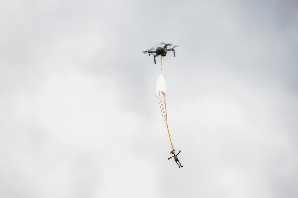 A drone carries a toy on a parachute up into the sky.