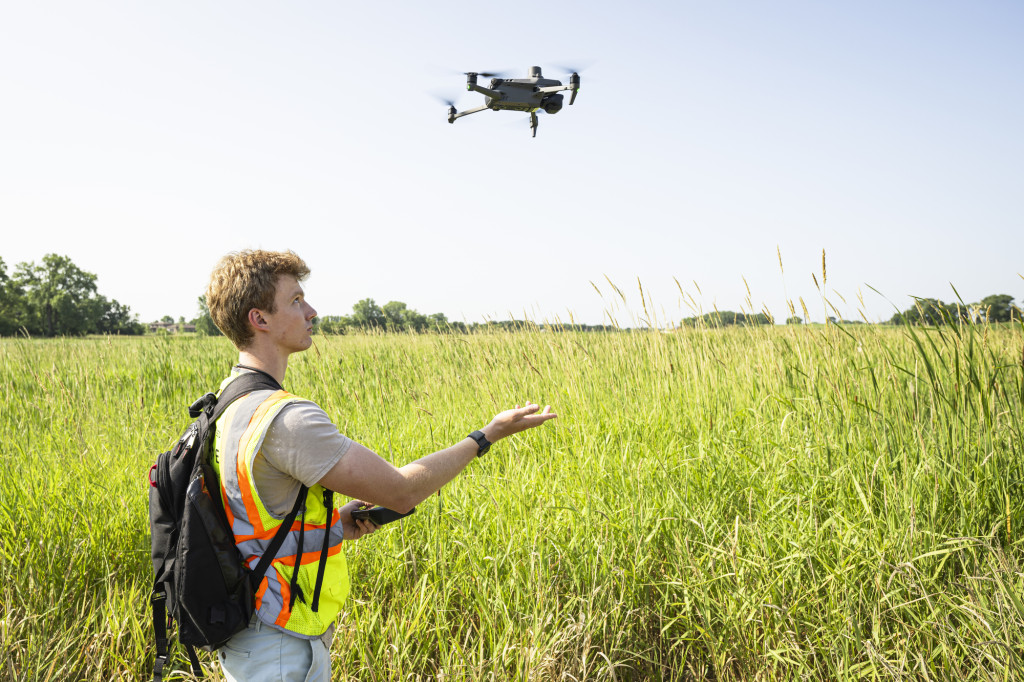 A man stands in a creek with a remote, as a drone flies overhead.