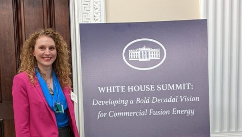 A woman in a pink blazer and black pants smiles as she stands next to a sign that reads White House Summit: Developing a Bold Decadal Vision for Commercial Fusion Energy. She is standing in a corridor of the White House.