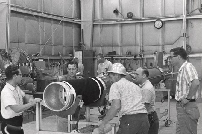 In a black and white photo from the 1960s, a group of men, some wearing hard hats, push a dolly carrying a long telescope tube. They're in an industrial warehouse setting.