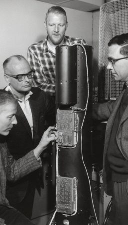 In a staged black and white photo from the 1960s, four men gather around a tall cylindrical piece of equipment.