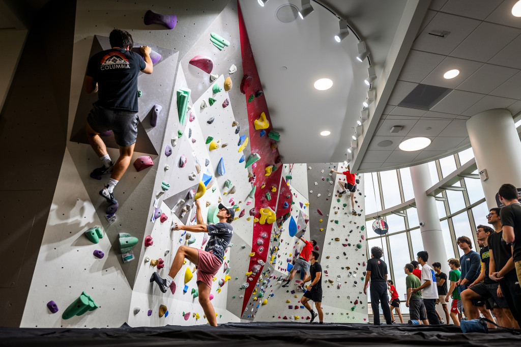 People climb on a bouldering wall, with ropes holding them up.