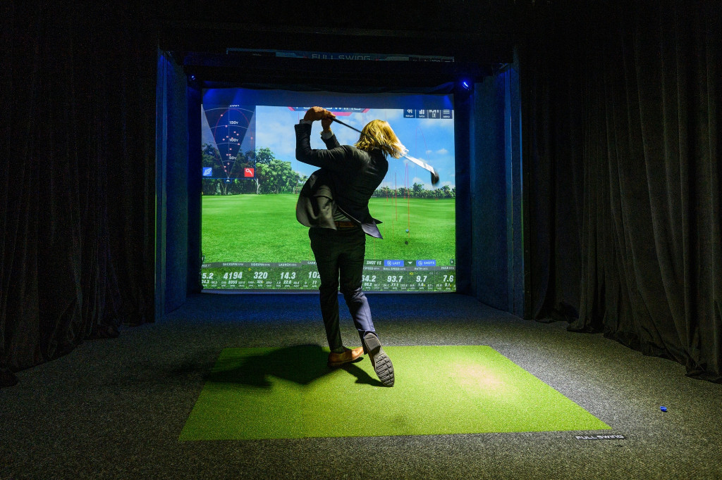 A person swings a golf club in front of a screen picturing a golf hole.