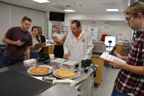 A group of people stand around a metal table and look at two cheese pizzas. They're in a laboratory setting.