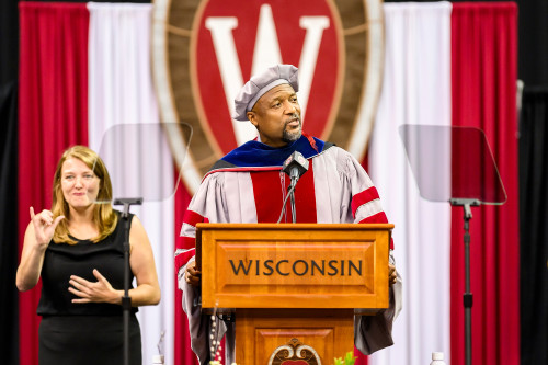 A man wearing academic robes speaks from a lectern.