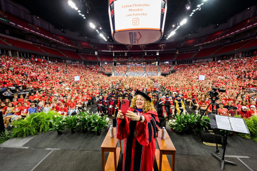 Chancellor Mnookin stands on stage wearing academic robes. She's taking a selfie with the thousands of people behind her filling the Kohl Center arena.