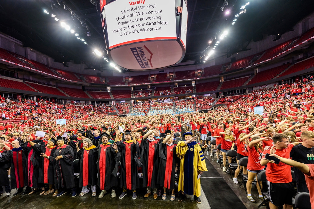 Thousands of people, most wearing red, some wearing academic robes, fill an arena. They're each waving one hand in the air as they sway in unison singing Varsity.