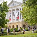 A view of Bascom Hall, a neoclassical building with white columns. Students walk on pathways in front of the building.