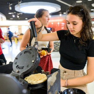 Olivia Cerro, a young woman in khaki shorts and a black T-shirt, makes a waffle with a waffle iron in a busy dining hall.