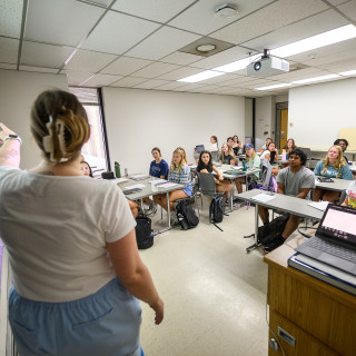 An instructor stands at the front of a classroom delivering a lecture to students seated at tables.
