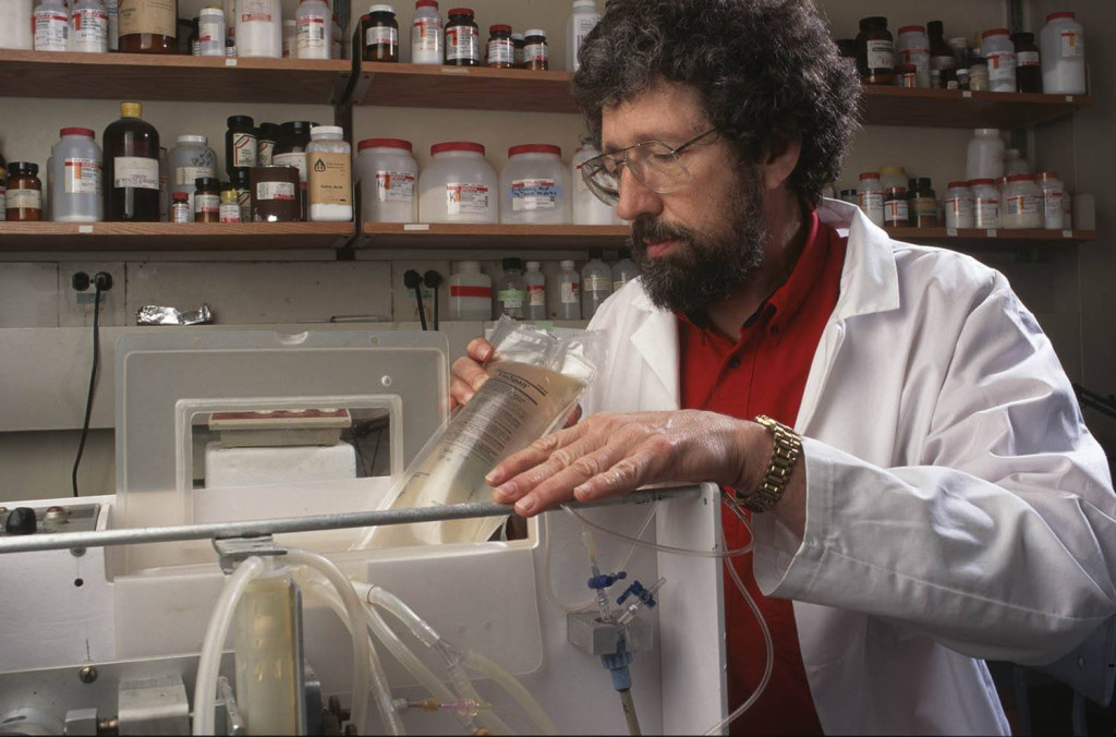 A an wearing a white lab coat over a red polo shirt holds a plastic bag filled with a liquid solution. He's looking into a piece of experimental equipment in a laboratory setting.