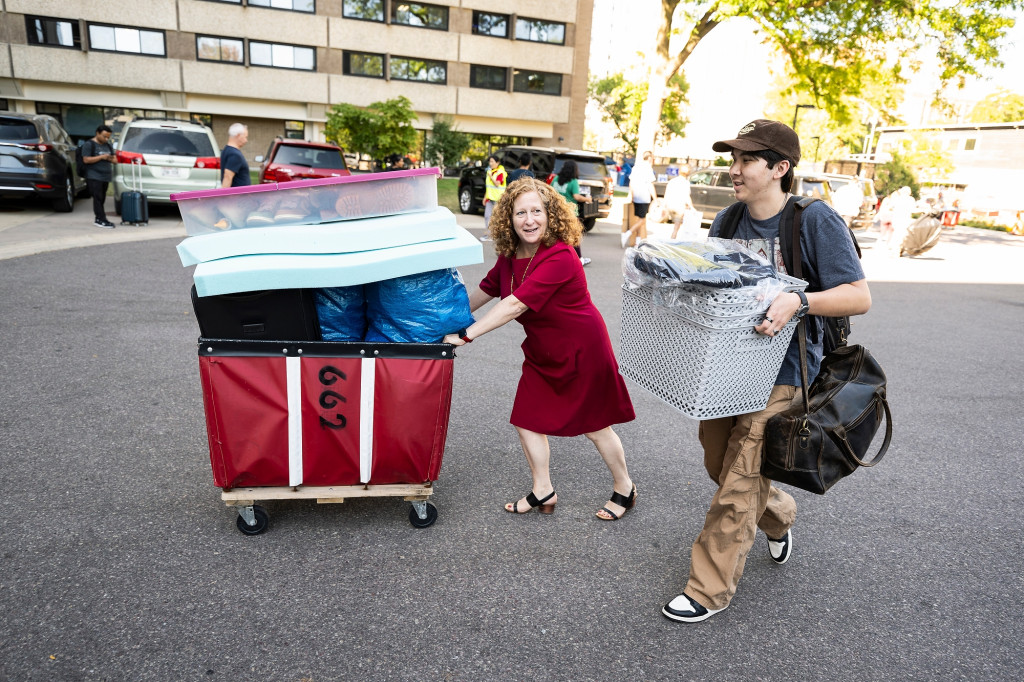 Chancellor Mnookin pushes a red moving cart filled with belongings across a parking lot. She's talking with a new student who has his hands full.