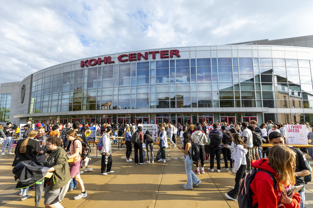 The shiny glass exterior of the Kohl Center is shown, with students walking around it.