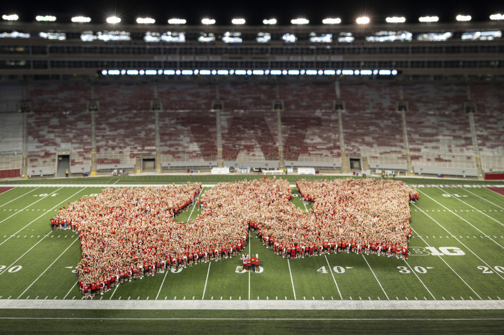 A crowd of people wearing red form a giant W on the 50 yard line in Camp Randall Stadium.