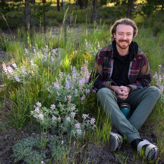 Timon Keller sits in a grassy area dotted by purple lupine flowers on the edge of a small stand of lodgepole pines.
