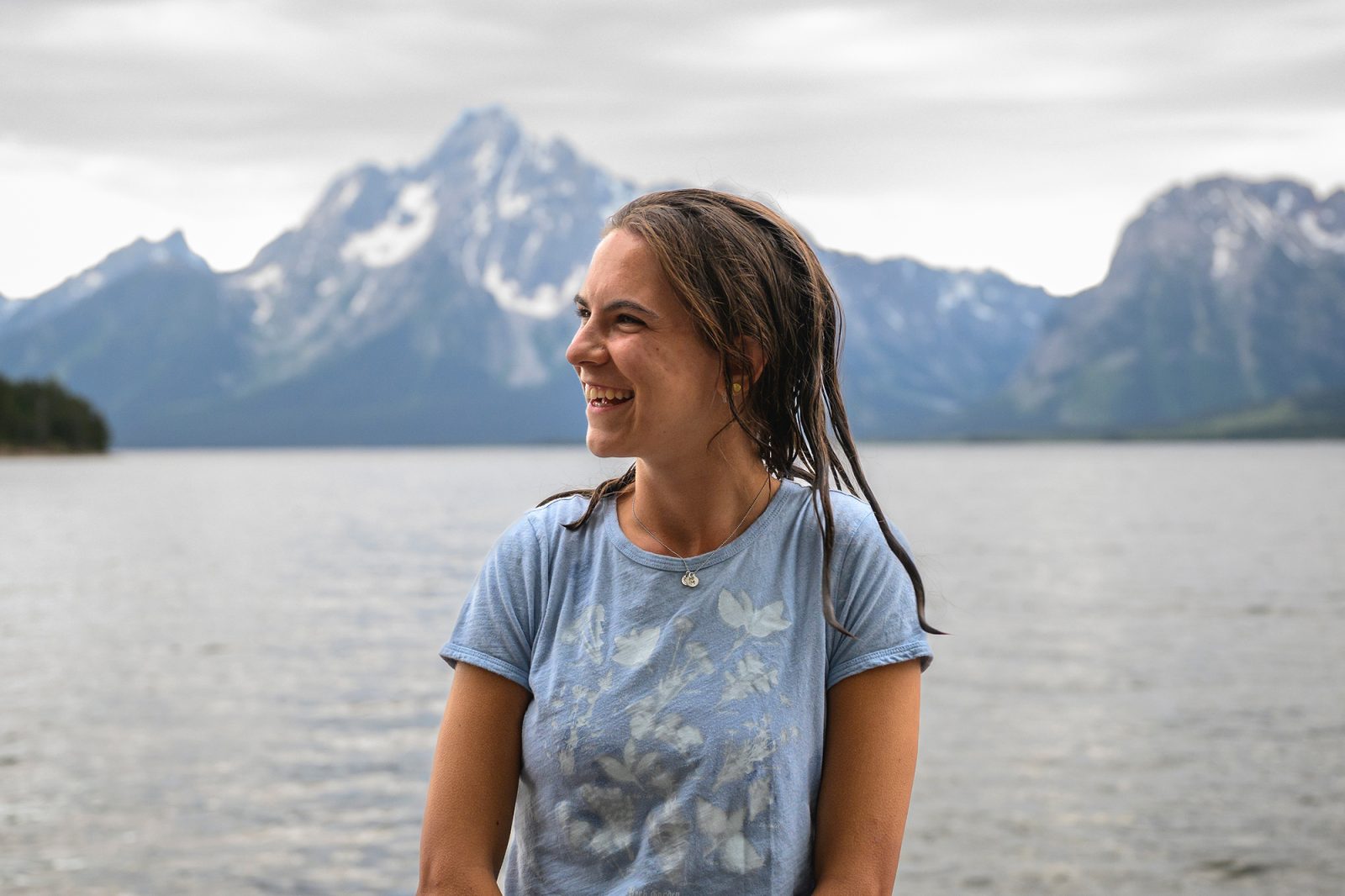 a woman smiling and looking off to the side in front while standing in front of snow capped mountains and a lake