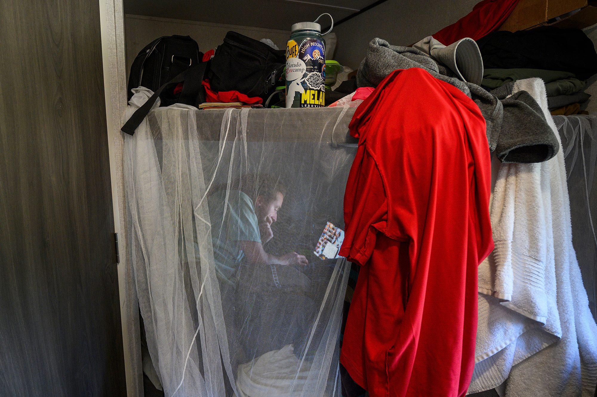Inside the team's RV, Timon Keller sits in the bottom bunk bed behind a gauzy translucent mosquito net that is hang from the top bunk which also stores water bottles, clothes and other personal belongings. His face is illuminated by the light of his laptop screen in the otherwise dark lower bunk.