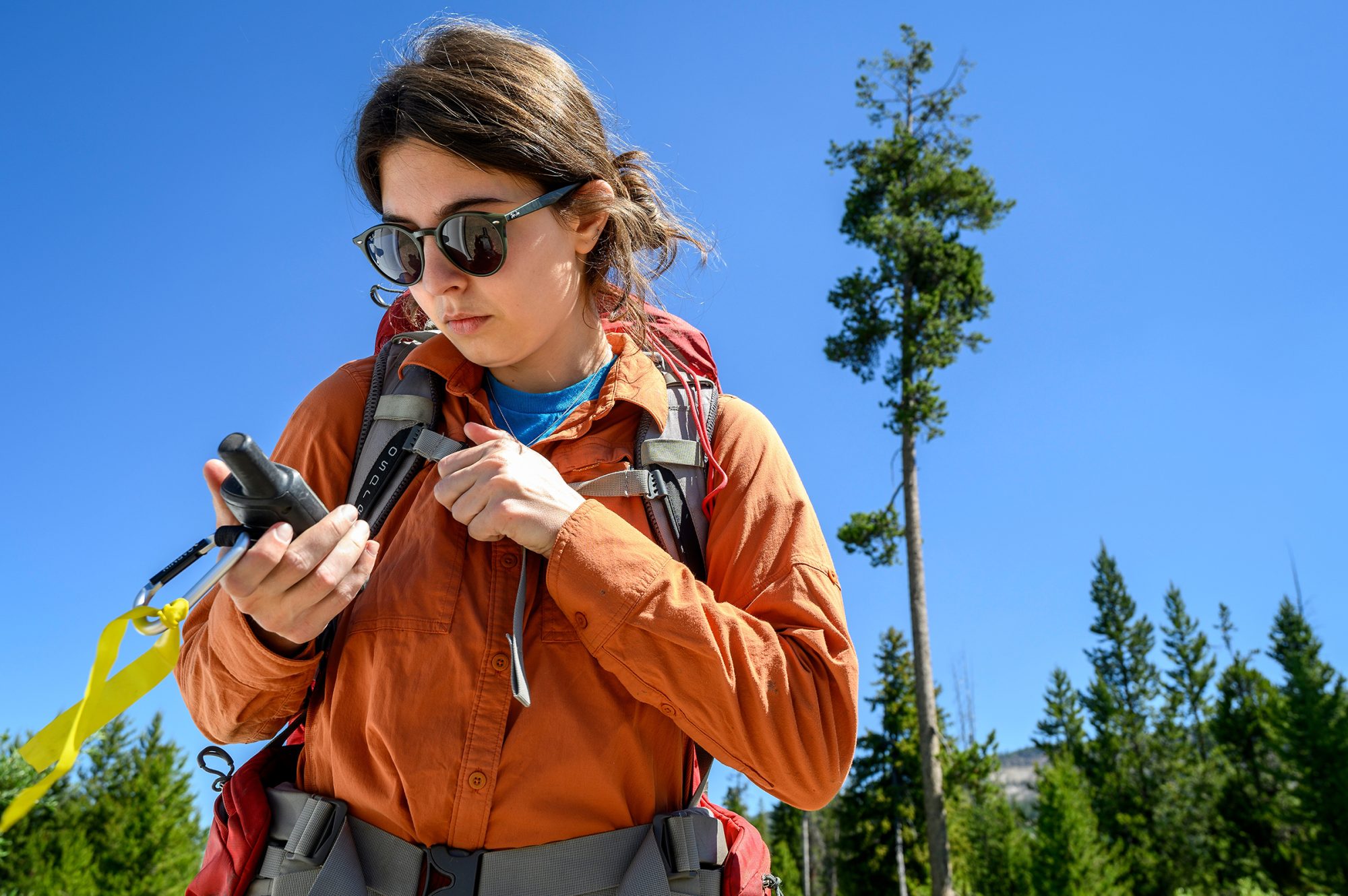 A woman in an orange shirt looks at a GPS unit while trees and blue sky show behind