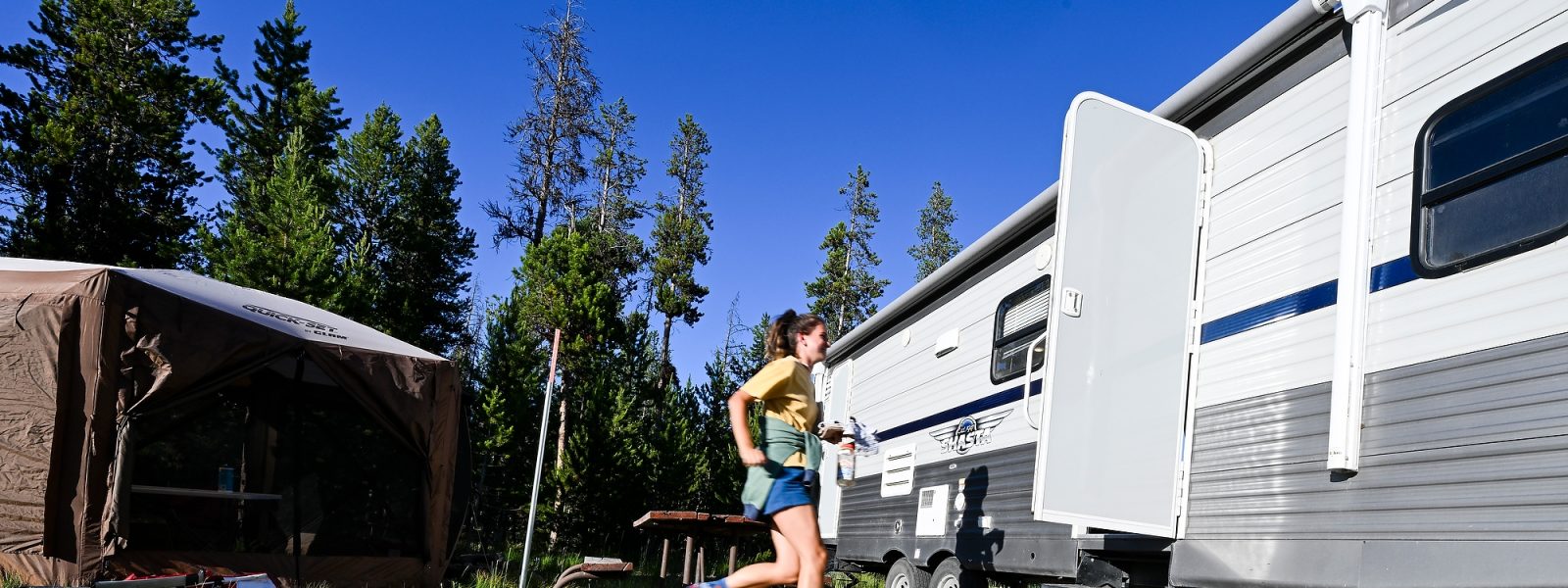Lucy McGuire runs through the middle of the frame from a large brown tent to the open door in the side of a white and grey RV parked at a campsite in Grand Teton National Park.