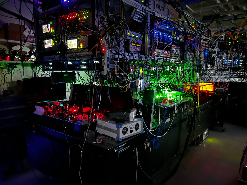 A photo of a dark room filled with computer hard drives and tangles of wires. Bright red, green, yellow and blue lights illuminate different blocks of equipment.