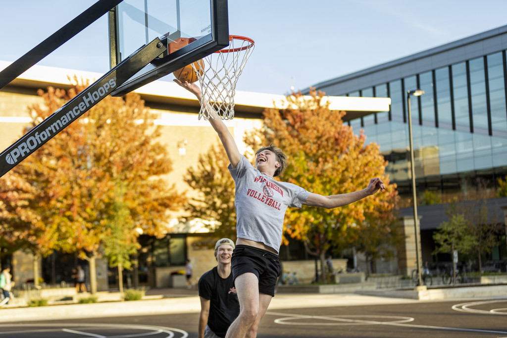 Outside a glass-walled campus building, a student wearing a Wisconsin T-shirt jumps for a layup shot in a one-on-one game of pickup basketball.