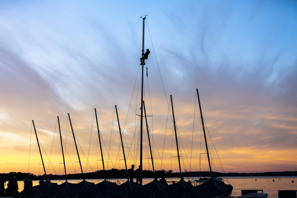 In silhouette against the setting sun, a person climbs the mast of a sailboat docked amid eight other sailboats.