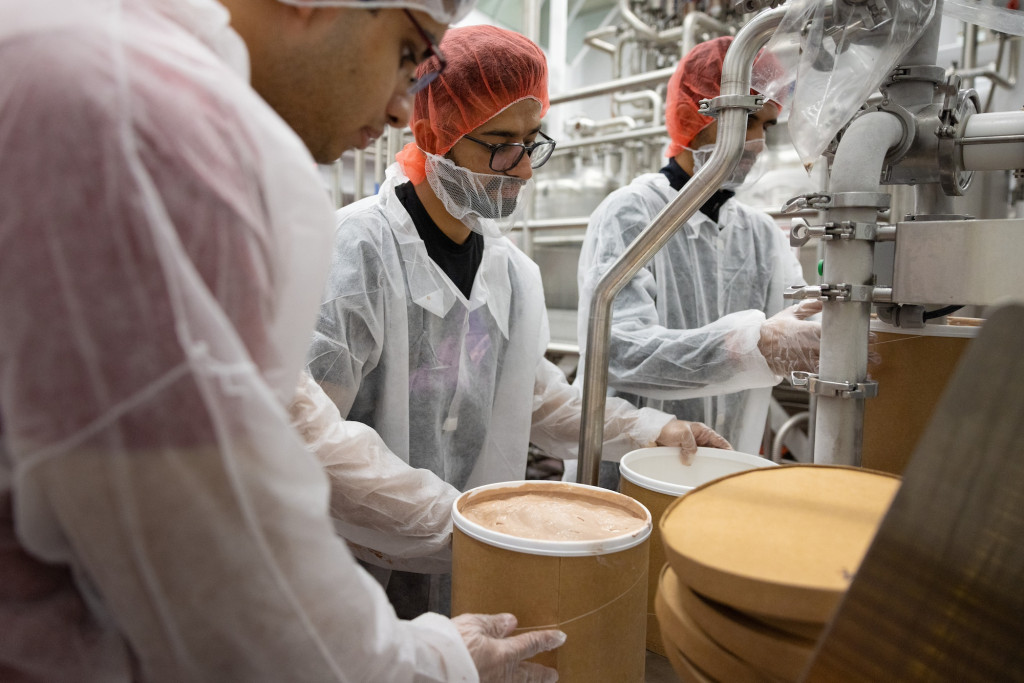 In an ice cream production facility, three people wearing hair nets and white food safety coveralls fill brown gallon containers with ice cream.