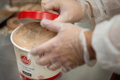 In a close-up phono, a person places a red lid on a container of ice cream labeled 175 S'more Years. The person is wearing clear food safety gloves.