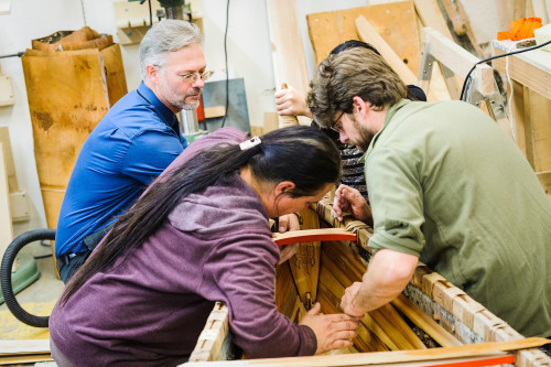 In a woodworking shop, four people talk to each other as they lean into the hull of a half-finished birchbark canoe.