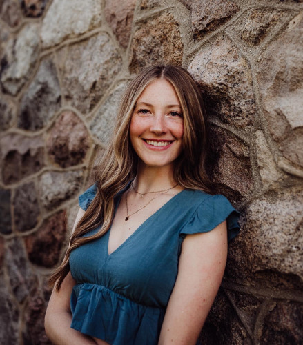 A portrait photo of Emily Hokanson. She is leaning against a stone wall and smiling to the camera.