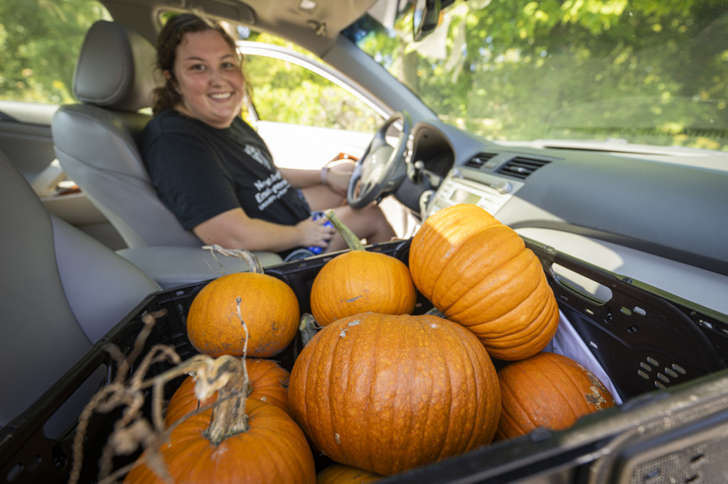 A woman in the driver's seat of a car smiles to the camera. The passenger seat is loaded with orange pumpkins.