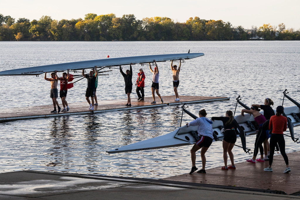 On the docks of Lake Mendota, two groups of students lift long crew watercraft overhead.