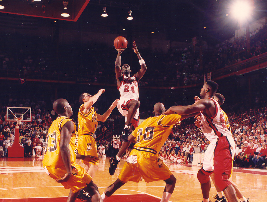 A man in a Wisconsin jersey takes a running jump shot as defenders look up at him.