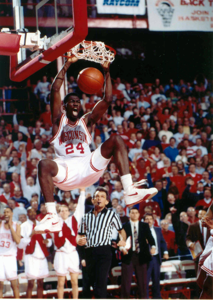 A man dunks a basketball as the crowd and the bench players cheer in the background.