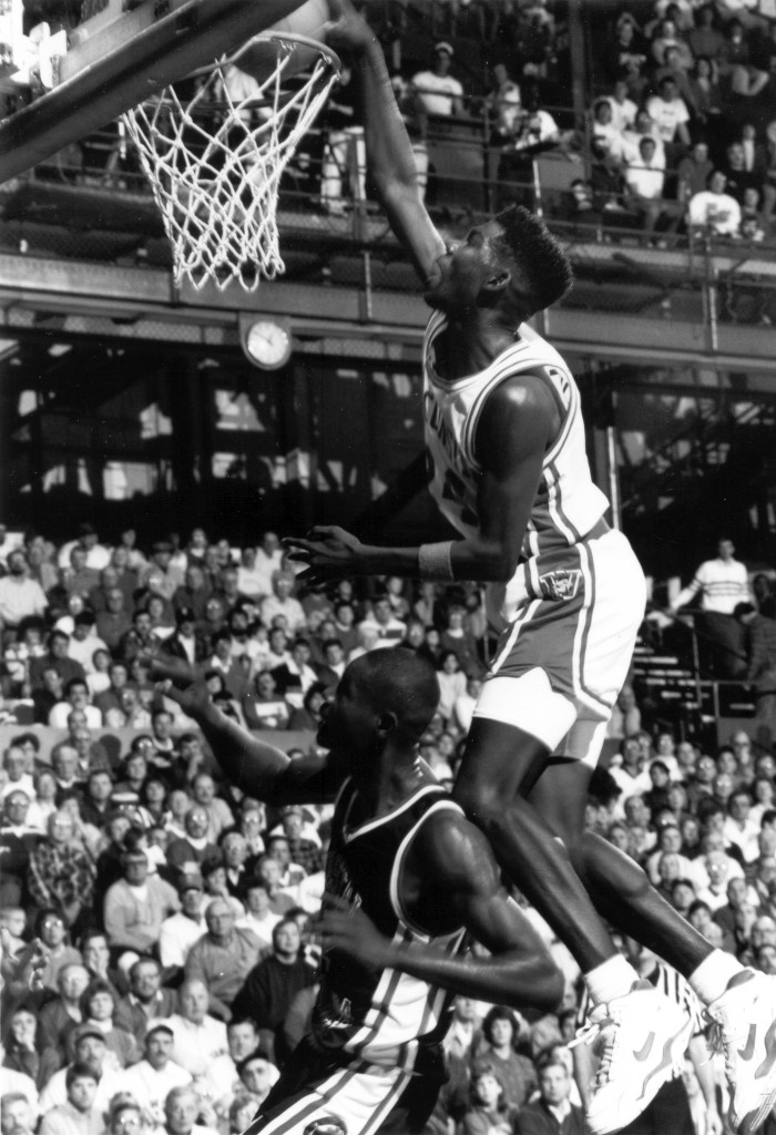 A photo of a man dunking over another man as the crowd watches.